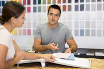 Obraz premium Male salesman of paintwork material department selects for female customer necessary color article of paint for concrete surfaces, walls in catalog