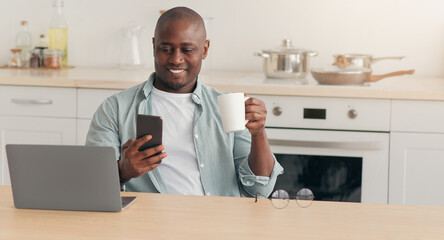 Morning coffee, news, chat in social networks, work and education at home. Smiling adult african american male disabled in wheelchair drinking, look at phone, at table with laptop in kitchen interior