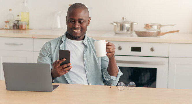 Morning coffee, news, chat in social networks, work and education at home. Smiling adult african american male disabled in wheelchair drinking, look at phone, at table with laptop in kitchen interior - Powered by Adobe