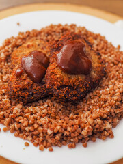 Simple tasty buckwheat and breaded chicken fillet with barbeque sauce meal on a white round plate.
