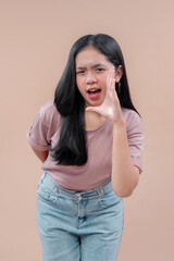 Asian woman in casual modern style leans forward with playful expression, holding hand near mouth as if shouting, wearing pastel t-shirt and denim jeans, showing confident youthful energy.