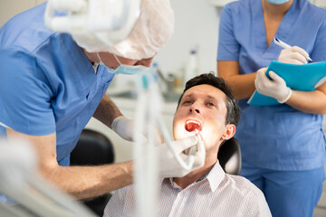 During work of senior male dentist and examination of male patients teeth, young woman nurse stands...