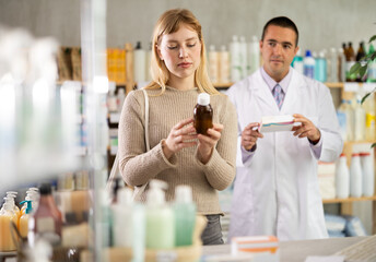 Girl studies composition of antitussive syrup, buys product in drug store. Checking for absence of GMOs, age restrictions and recommendations for use. Man pharmacist in background offers similar