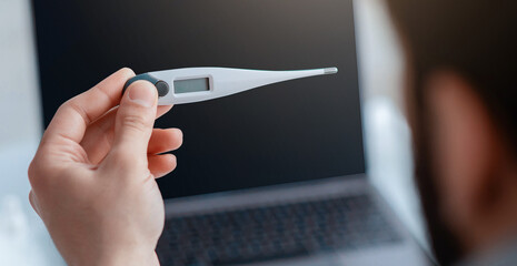 Sick during work from home. Man holding digital thermometer in front of laptop computer, selective...