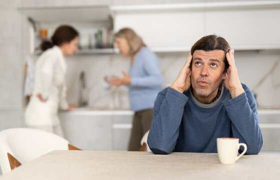 Frustrated man sitting in kitchen during quarrel between his wife and elderly mother. Complicated family relationships, daughter-in-law and mother-in-law yell at each other - Powered by Adobe