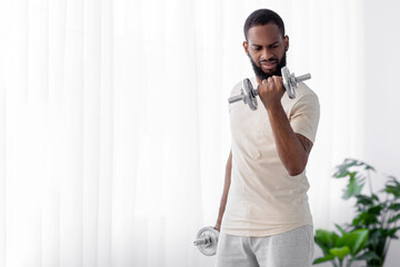 Strong young black male with beard in white sportswear raises dumbbell in room interior on window...