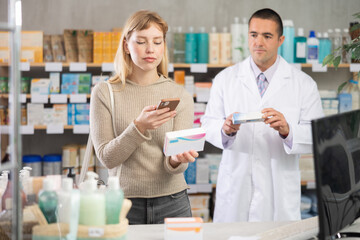 Young woman in pharmacy scans QR code on probiotic pills product, studies item in detail, collects information online. Client learn reviews and recommendations for use. Man pharmacist in background.