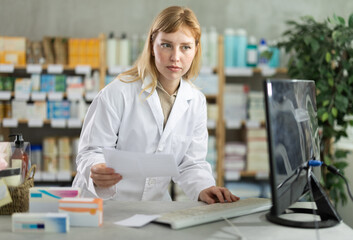 Qualified female pharmacist looks at a medical prescription from a customer and checks the...