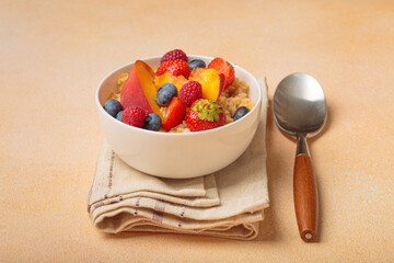 Oatmeal porridge with fresh blueberries, strawberries, raspberries, and peach slices is served in a white bowl on a napkin with a spoon for a healthy and nutritious breakfast