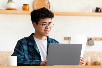 Work, paperwork and remote education at home during covid-19 pandemic. Smiling asian millennial guy employer in glasses typing on laptop, looks documents on table with coffee in boho kitchen interior