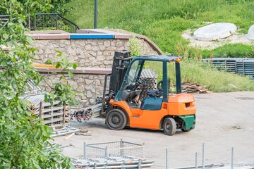 An orange industrial forklift parked in an outdoor storage or construction area, adjacent to a rock formation and trees The ground is uneven with debris scattered, under soft diffused lighting © Mister