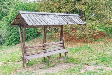 Tranquil outdoor setting, rustic wooden bench under a slatted roof, lush greenery, natural...