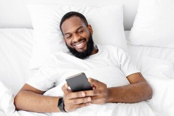 Smiling young black bearded guy lies on white bed in bedroom, checks application and social networks on smartphone. Covid-19 pandemic, chat, new morning news and positive facial expression, top view