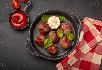 Fried meatballs in a cast iron pan, surrounded by basil, aromatic garlic, and tomato sauce on black...