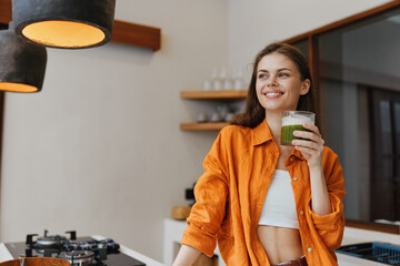 Smiling young woman in an orange shirt holding a green smoothie in a modern kitchen, showcasing...