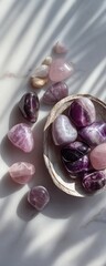 A shallow, light beige bowl filled with various tumbled, heart-shaped crystals in shades of lavender, purple, and pink sits on a light gray marble surface, dappled with sunlight
