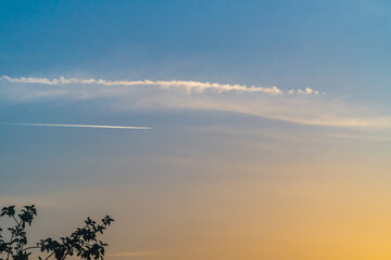 A sunset cloudless blue sky with a haze that is illuminated by yellow sunlight and streaks of traces left by the flight of planes, another flies in a northerly direction