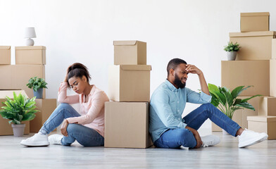 Tired upset young black husband and wife among cardboard boxes with things and plants in empty room...