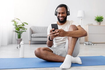 Satisfied millennial black guy in headphones sits on floor on mat in room interior, typing on phone...