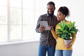 Glad young african american couple looking in tablet, wife holding plant, standing near window in...