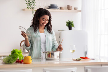 Healthy eat, food blog and cook dinner at home. Quarantine, homemade dish. Millennial pretty african american female make soup in modern white kitchen interior with bright vegetables, empty space