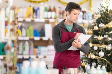 Man seller, using paper report make inventory in household chemistry store. Employee plans to replenish restock household stores inventory