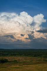Massive cumulonimbus cloud dominates the sky, casting a dramatic presence over a serene landscape of coniferous forests and open fields, hinting at an approaching storm