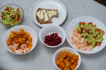 Table is set with a variety of healthy lunch options. Including shrimp. A fresh salad. A slice of buttered bread. Cooked pumpkin. And a bowl of red berries. Offering a balanced and nutritious meal