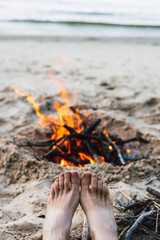 A person's feet relaxing by a bonfire on a sandy beach. Bare feet and a campfire on a sandy beach, with the ocean in the background, creating a relaxing outdoor scene. Female tourist Slow living scene