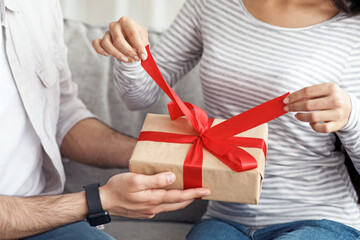 Holiday at home, new normal, covid-19 and birthday, anniversary and valentine's day. Millennial arab lady opens gift with red ribbon from her husband on sofa in living room interior, close up, cropped