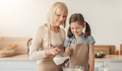 Learning How To Cook. Portrait of cheerful little girl in apron kneading dough using whisk, smiling nanny pouring milk in glass bowl, family cooking together trying new recipe, making sweet cake