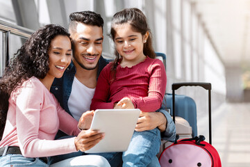 Beautiful young middle eastern family using digital tablet at airport hall, happy arabic parents...