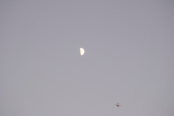 A striking half moon is visible high in a soft, twilight sky, with the small silhouette of a flying airplane near the bottom. This minimal image beautifully combines themes of night travel, space, and