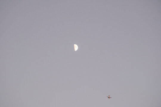 A striking half moon is visible high in a soft, twilight sky, with the small silhouette of a flying airplane near the bottom. This minimal image beautifully combines themes of night travel, space, and