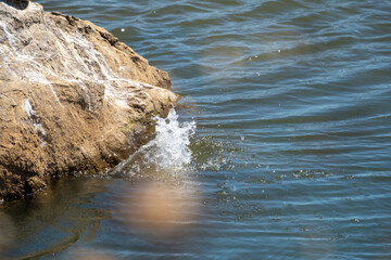 Close-up shot of water splashing dramatically against a large, textured rock formation at the edge of a reservoir. Focus on the dynamic motion, clear spray, and the contrasting textures of water and s