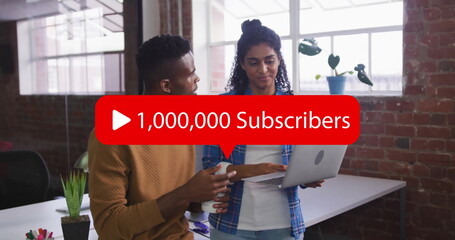 Collaborating coworkers pointing at silver laptop with red subscriber overlay in office with mug
