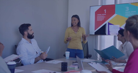 Presenting professional in mustard blouse gesturing toward display screen at boardroom with laptops