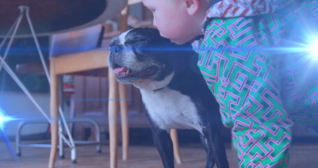 Kneeling toddler boy in patterned hoodie kissing dog beside chair in living room, copy space