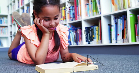 Lying child wearing peach top, purple shorts reading open book on library carpet, with bookshelves
