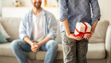 Boy greeting his young dad with Father's Day, hiding gift box behind his back, selective focus. Dad...