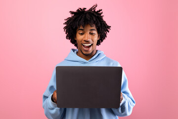 Happy black teenager looking at laptop screen with smile, watching interesting web content on pink studio background. Cool African American teen guy communicating to friend online, browsing web