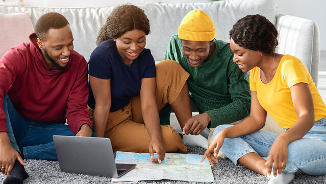 Smiling black friends sitting on floor at home, planning summer vacation, using map and laptop. Group of african american young men and women travellers booking flights and hotels