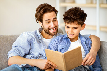 Useful time at home. Father and son reading book and smiling, resting on comfy sofa in living room....