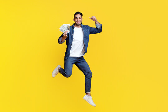 Saving money, gambling, gaming, trading, loan concept. Positive middle-eastern guy holding bunch of money and raising fist up, won lottery, yellow studio background, full length, copy space