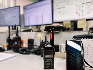 A desk in a control room with multiple walkie-talkies, computer monitors displaying data, and a smartphone. Essential equipment for security, communication, and operational monitoring.