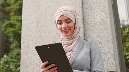 A young business woman wearing a light-colored hijab smiles while browsing on her tablet outdoors. She stands near a concrete pillar, surrounded by greenery, reflecting a modern lifestyle. - Powered by Adobe