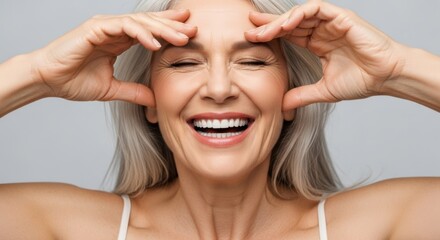 An ecstatic senior woman with gray hair laughs while stretching her facial skin