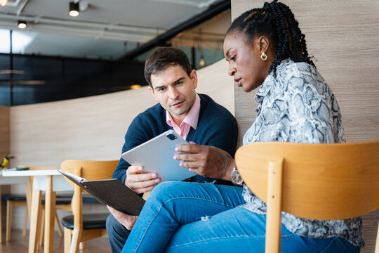 A diverse business team collaborating on a tablet in a modern office or cafe setting, Two colleagues, a man and a woman, meeting and reviewing a digital document and notebook together