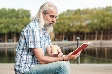 Senior man reading a book and drinking coffee by the river