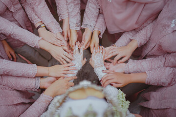 Bridesmaids in matching lace dresses gather around the bride, placing their hands over hers adorned with elegant white lace gloves in a heartfelt wedding moment.